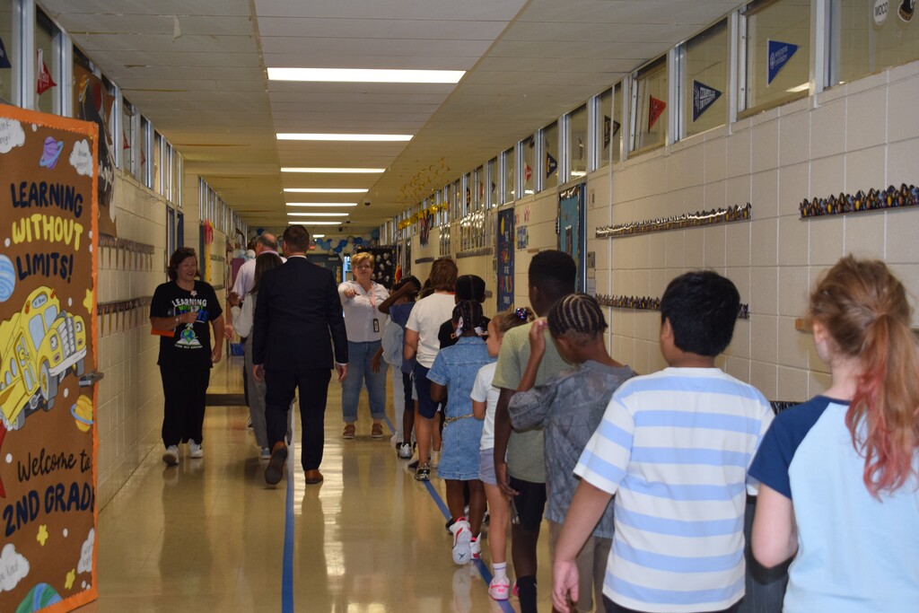 Crowded school hallway with students standing at lockers and walking through the corridor, colorful posters on the walls.