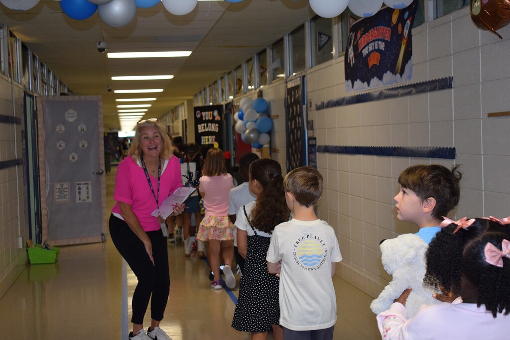 A group of children and a teacher walking through a decorated school hallway with balloons and banners.