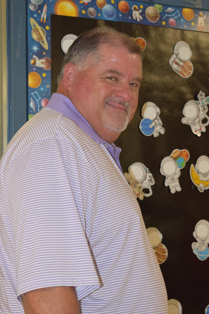 A middle-aged man with gray hair and a beard, wearing a striped shirt, standing in front of a playful background with colorful space-themed decor.