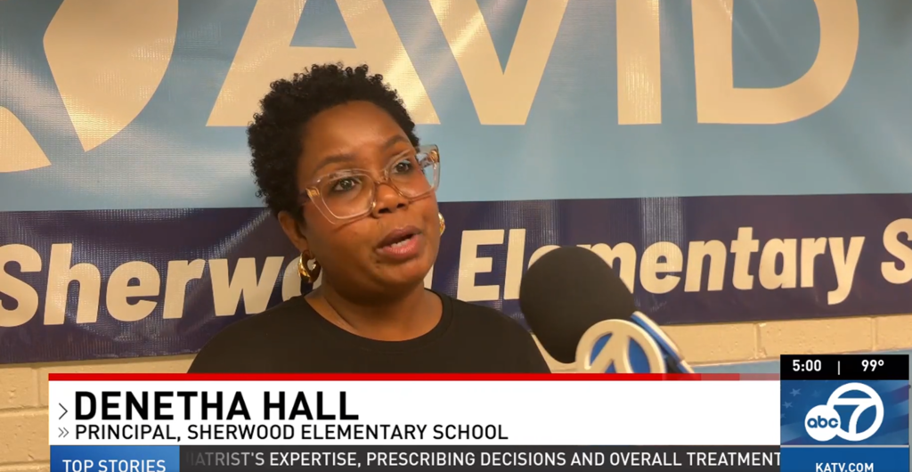 The image shows a middle-aged woman with glasses and curly dark hair, standing in front of a banner for Sherwood Elementary School, where she is the principal.