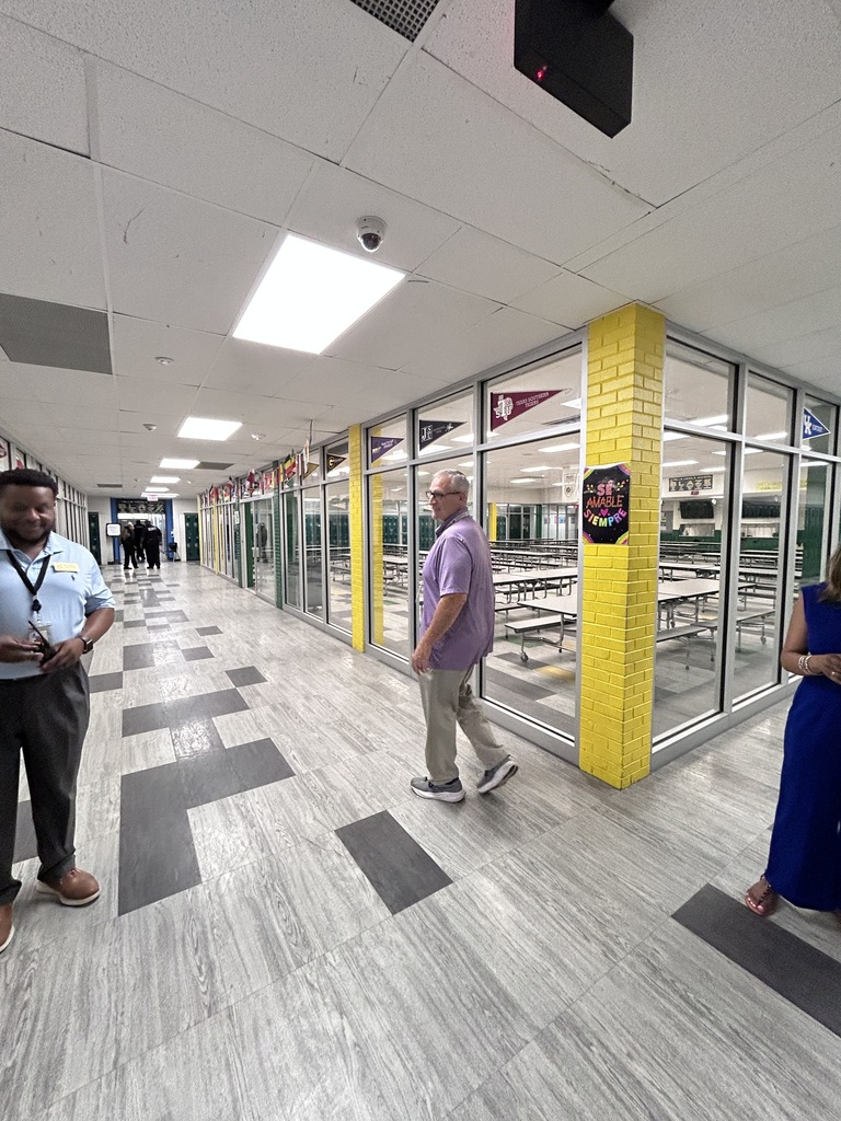 An interior hallway with tile flooring and glass-enclosed rooms. Two people walking in the hallway. Large yellow column and various signs visible.