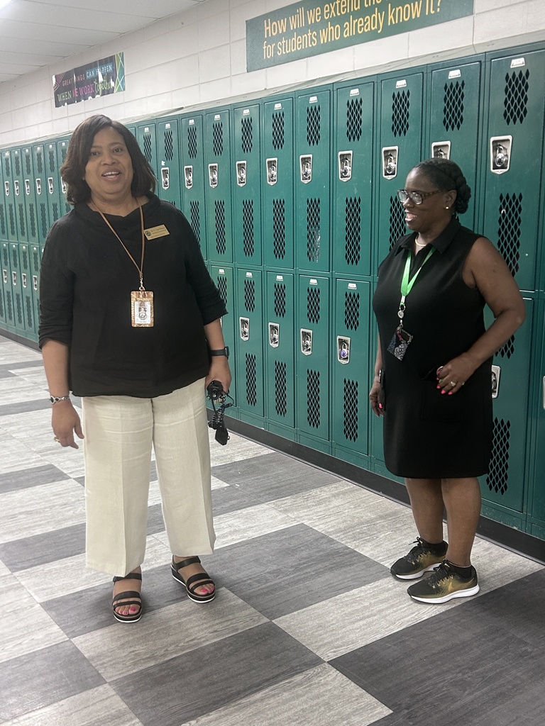 Two women standing in a school hallway near lockers with a sign that says "How will we extend our...for students who already know it?"