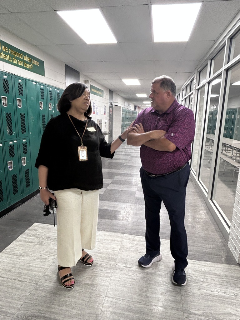 Two people conversing in a school hallway; a woman wearing a name tag stands speaking with a man in casual clothing.
