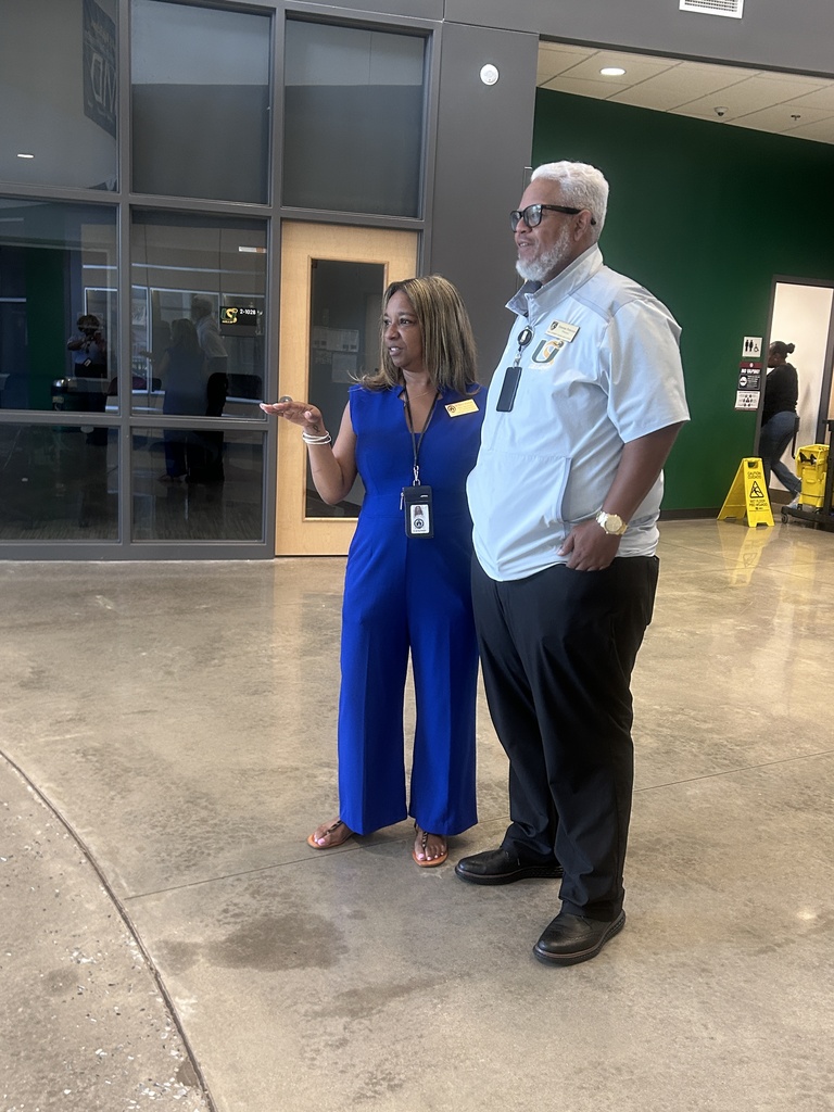 Two people in uniforms, a woman in a blue jumpsuit and a man in a white shirt, standing in a lobby with a glass wall.