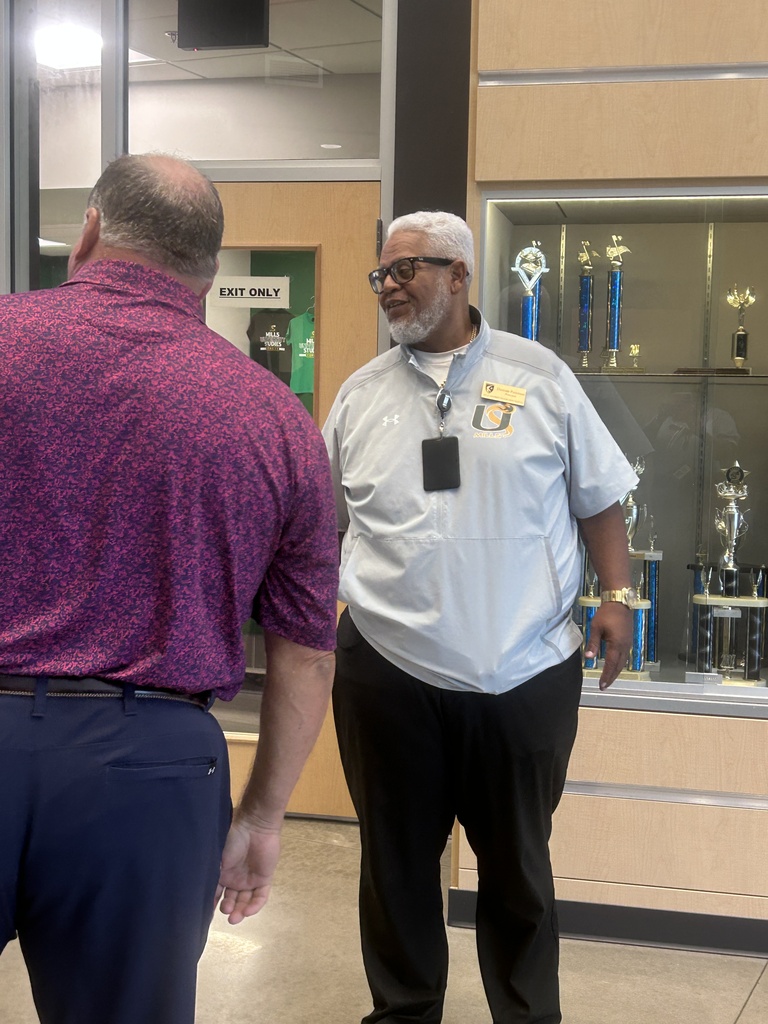 Two men in conversation in a school hallway, with one wearing a white shirt and the other wearing a purple jacket.