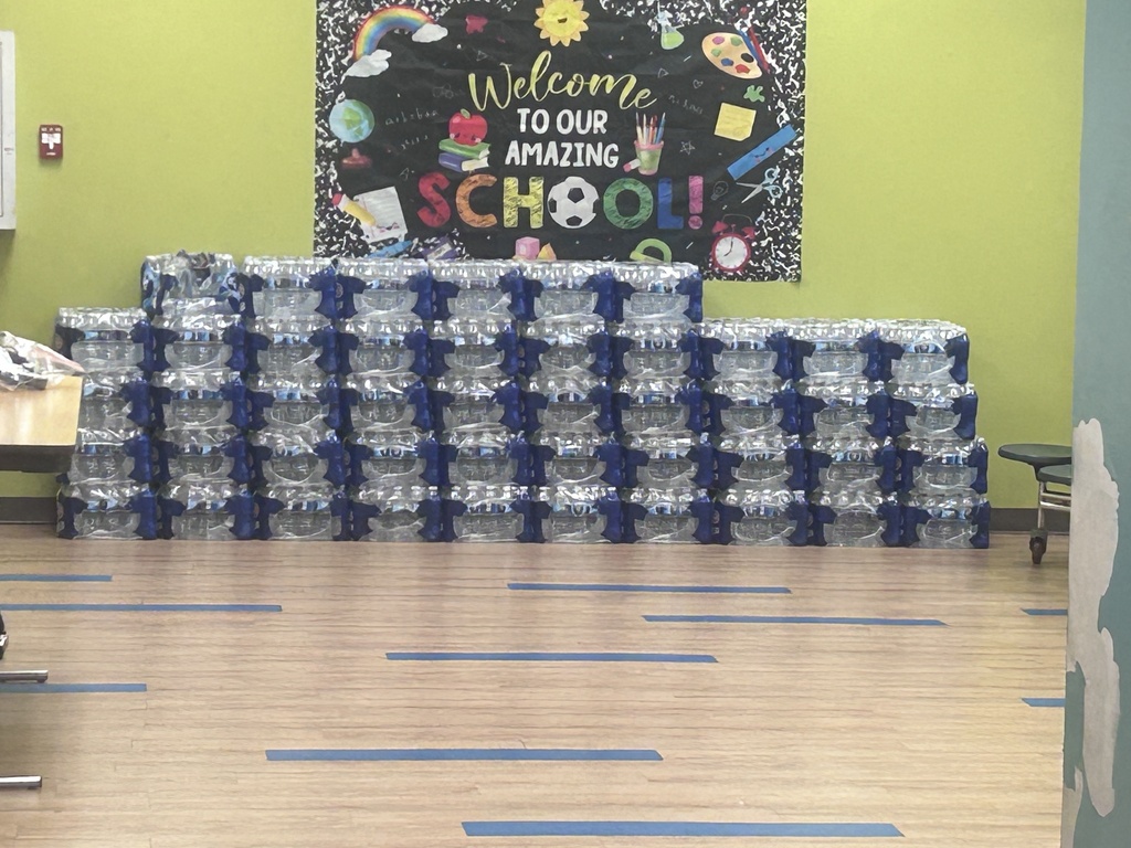Stacks of plastic water bottles on the floor of a school cafeteria with a colorful bulletin board in the background.