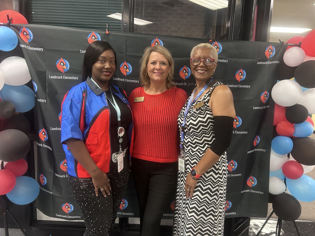 Three women standing in front of a backdrop with the logo of a cardinal. The women are smiling and wearing colorful outfits.