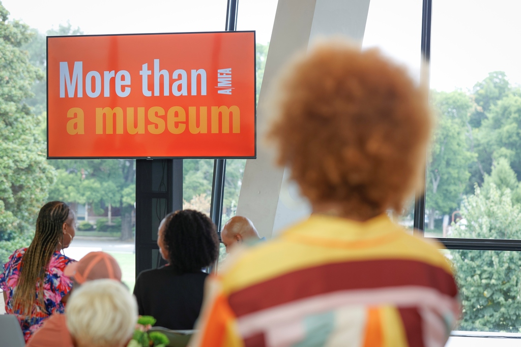 A screen in the background says More than a Museum AMFA. People in the foreground are looking toward it.