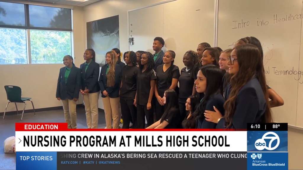 A group of Mills High students pose in a classroom, some wearing scrubs and others in khaki pants with green shirts and navy blazers. A whiteboard behind them reads “Intro into Healthcare” and “Medical Terminology,” shown during a KATV education segment.
