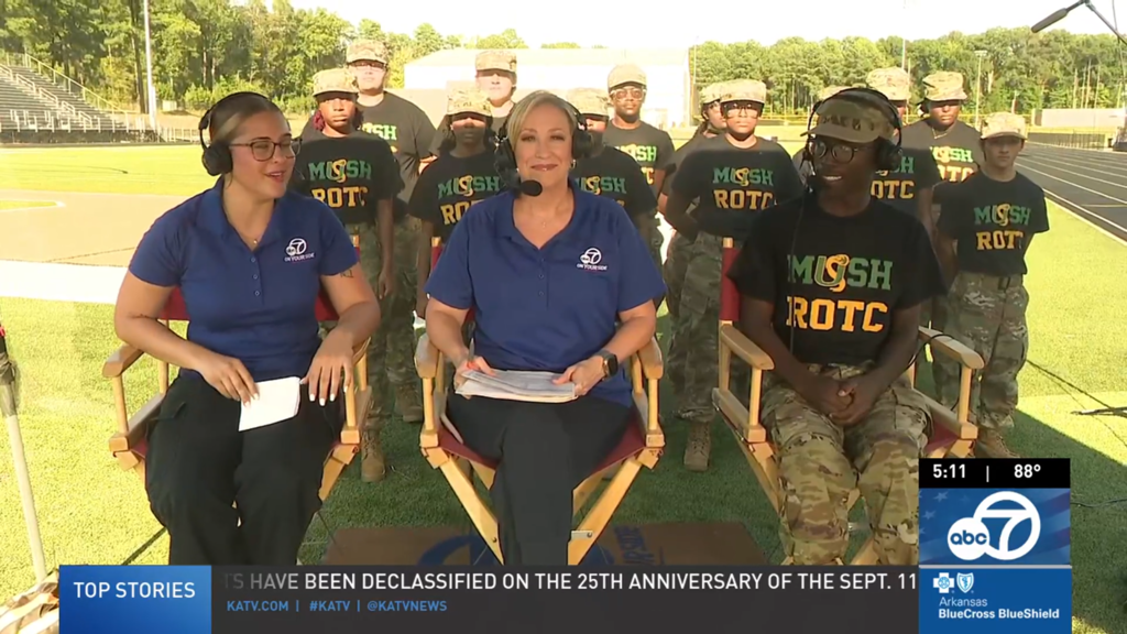 A Mills High ROTC student sits on a sports field with two ABC7 reporters during an outdoor interview. Behind them, additional ROTC students stand in formation wearing matching shirts and camouflage pants.