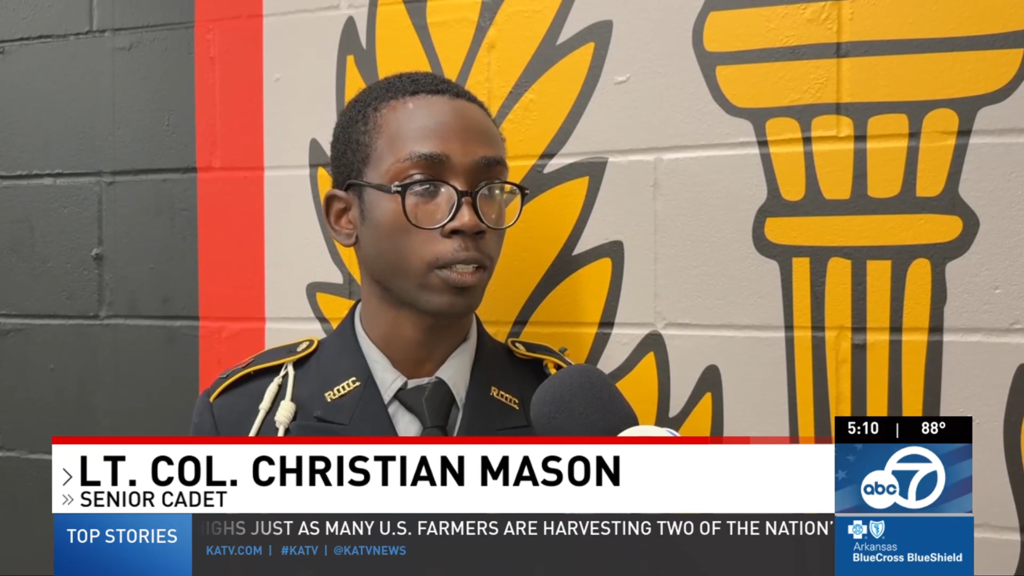 Senior Cadet Lieutenant Colonel Christian Mason stands in uniform in front of a painted mural while being interviewed on camera. A KATV lower‑third identifies him as a senior cadet during a news segment.