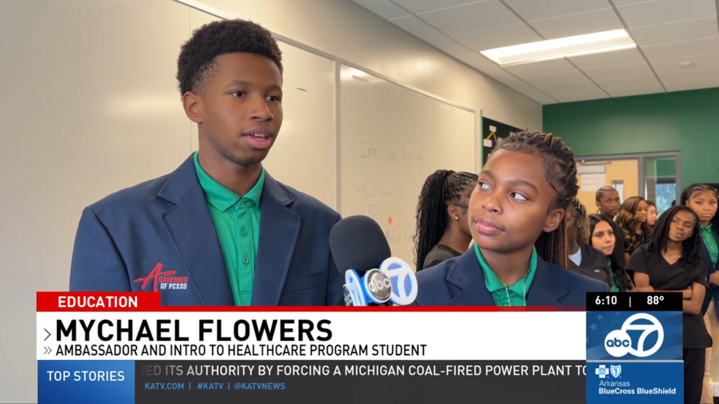 Healthcare ambassador Mychael Flowers speaks into a reporter’s microphone while another student stands beside him. Both wear green collared shirts and navy blazers with the Academies of PCSSD logo, with classmates visible behind them in a KATV education segment.