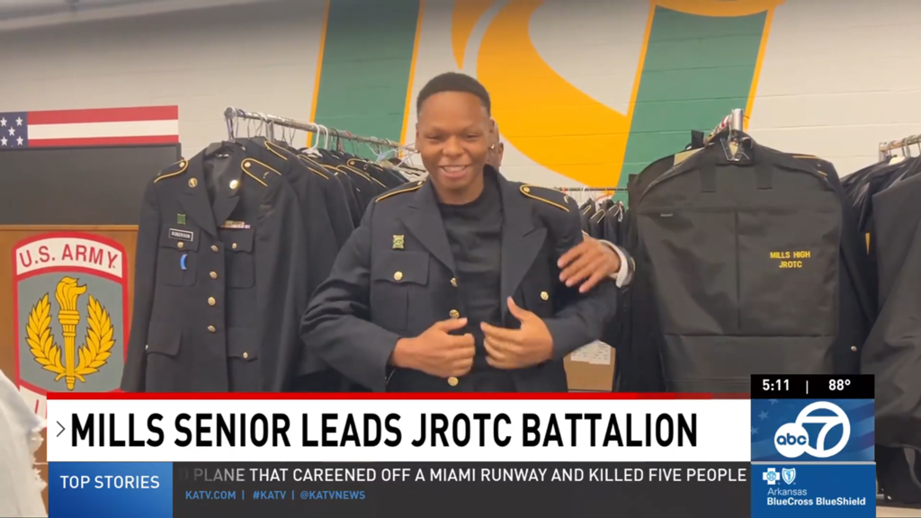 A Mills High JROTC student stands in a uniform room wearing a dark formal jacket, surrounded by racks of military‑style uniforms. A U.S. Army emblem and a garment bag labeled “MILLS HIGH JROTC” appear behind him, shown in a KATV news segment titled “MILLS SENIOR LEADS JROTC BATTALION.”