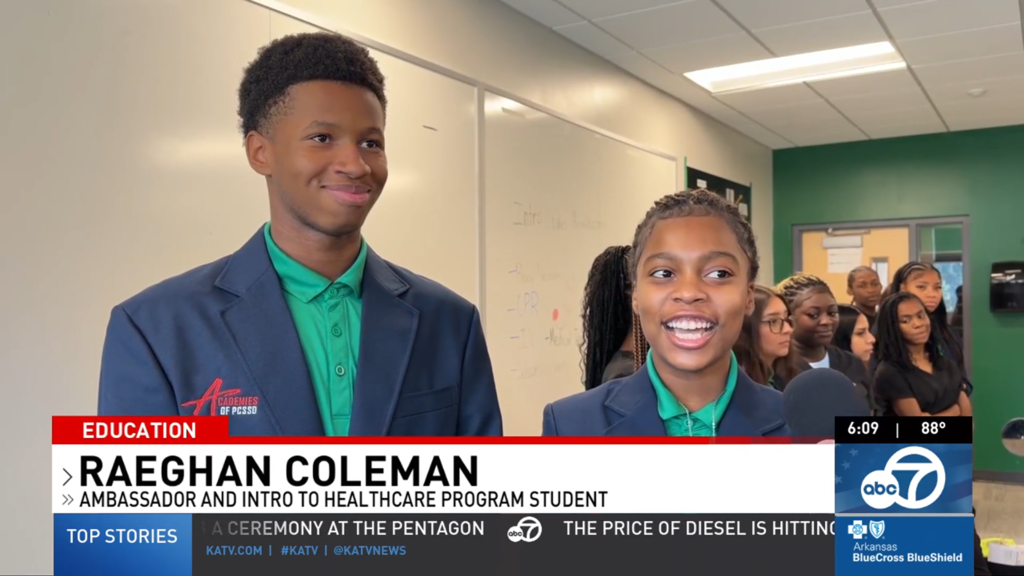 Healthcare ambassador Raeghan Coleman speaks into a microphone while another student stands beside her. Both wear green collared shirts and navy blazers with the Academies of PCSSD logo, with additional students lined up behind them in a classroom during a KATV education segment.