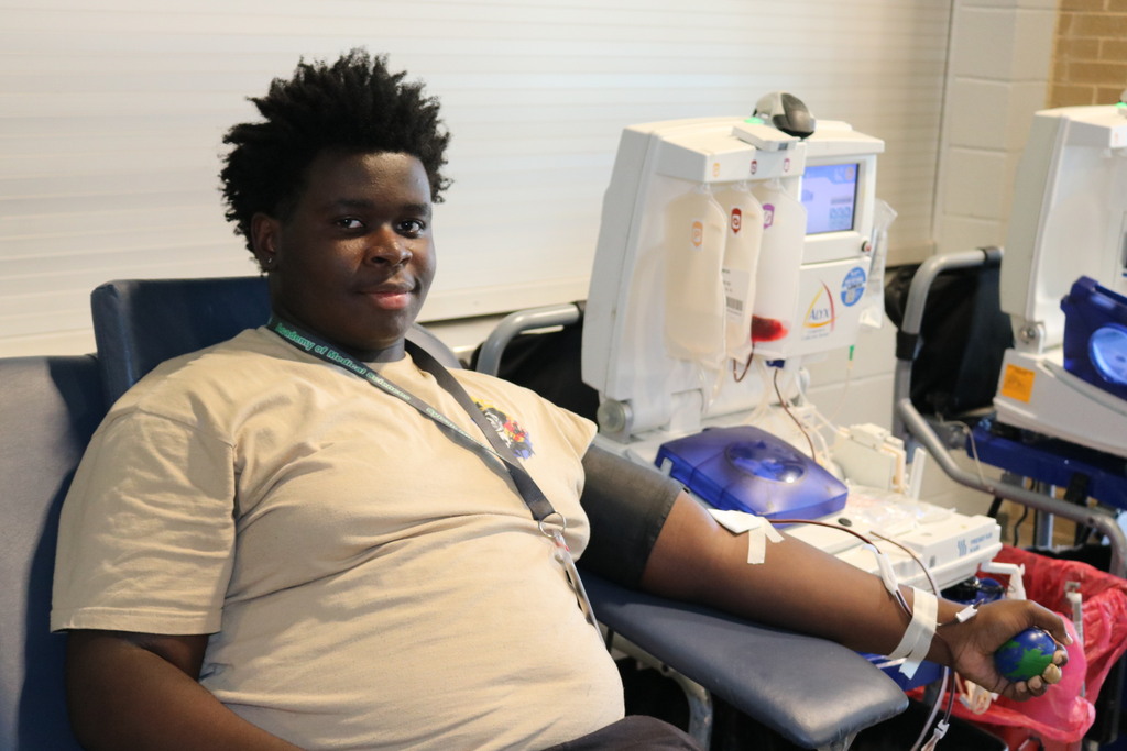 A student sits in a medical donation chair with a needle in their arm connected to tubing and a collection machine. Several attached bags contain fluids. The student squeezes a small green ball and wears a beige shirt and lanyard.