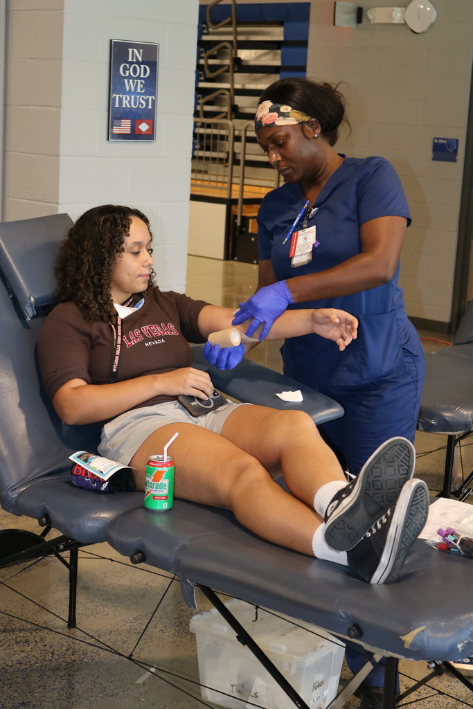 A person reclines on a medical cot while a staff member in scrubs applies a bandage to their arm. A drink and snack sit beside the donor. Medical supplies and a sign on the wall are visible in the background.