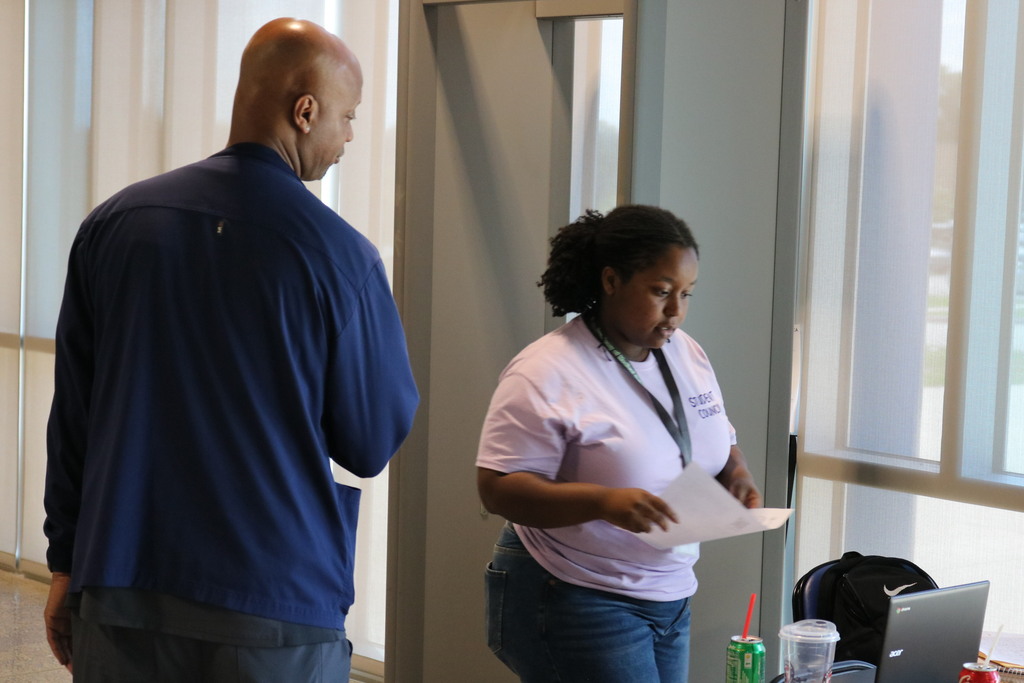 Two individuals stand indoors near a window. One wears a dark jacket facing away; the other wears a light purple “STUDENT COUNCIL” shirt and holds a paper. A laptop, backpack, drink cup, and soda can sit on the table beside them.
