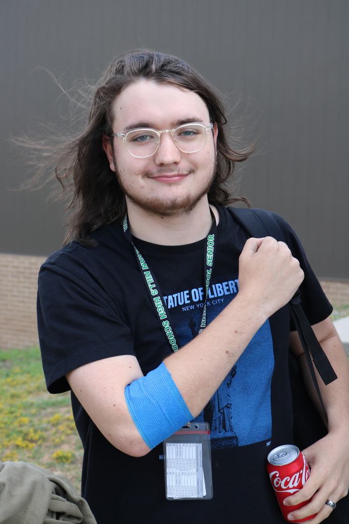 A student stands outside near a building wall wearing glasses, a black T‑shirt with a blue Statue of Liberty design, and a green lanyard. A blue bandage is wrapped around the student’s right arm, and they hold a red soda can in their left hand. Grass and small yellow flowers are visible behind them.