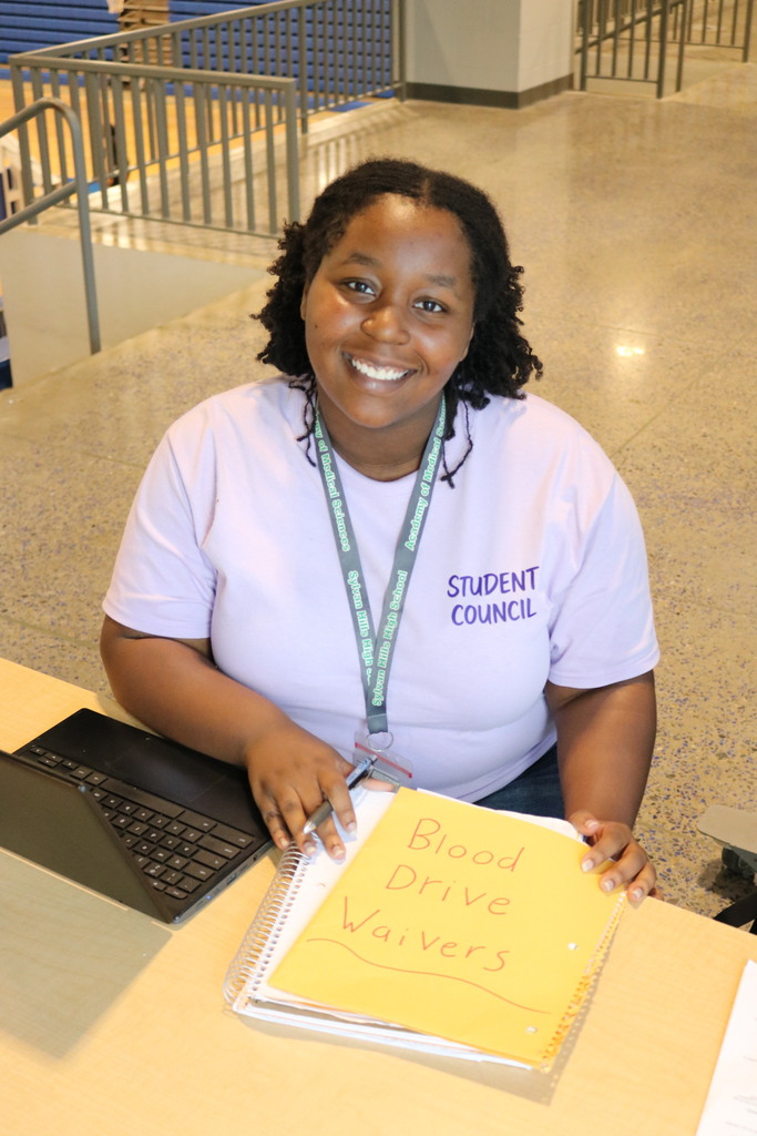 A student wearing a light purple “STUDENT COUNCIL” shirt sits at a table with a laptop and a yellow folder labeled “Blood Drive Waivers.” Metal railings and tiled flooring are visible in the background.