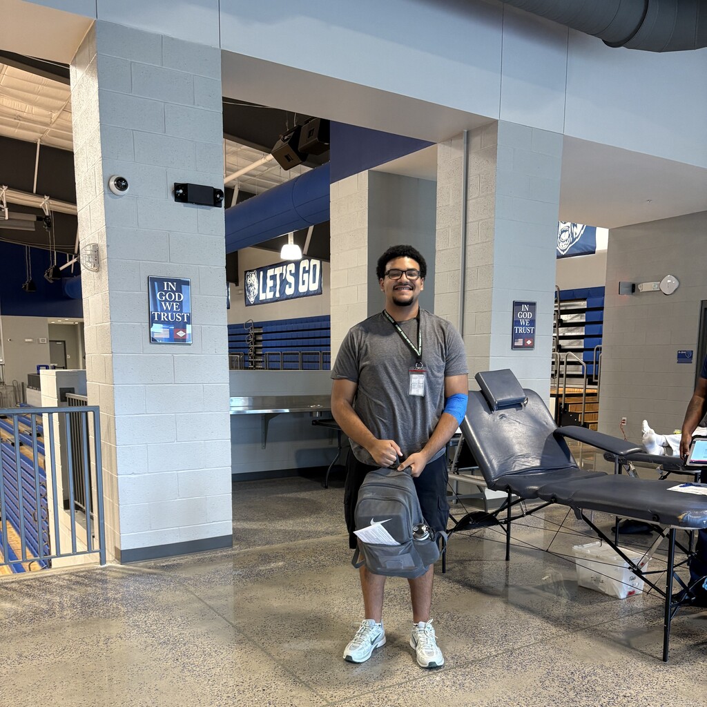 A student stands in a gym holding a gray backpack, wearing a gray shirt, black shorts, and white sneakers. A blue bandage is on their arm. Behind them are reclining donation chairs, medical supplies, and wall signage.