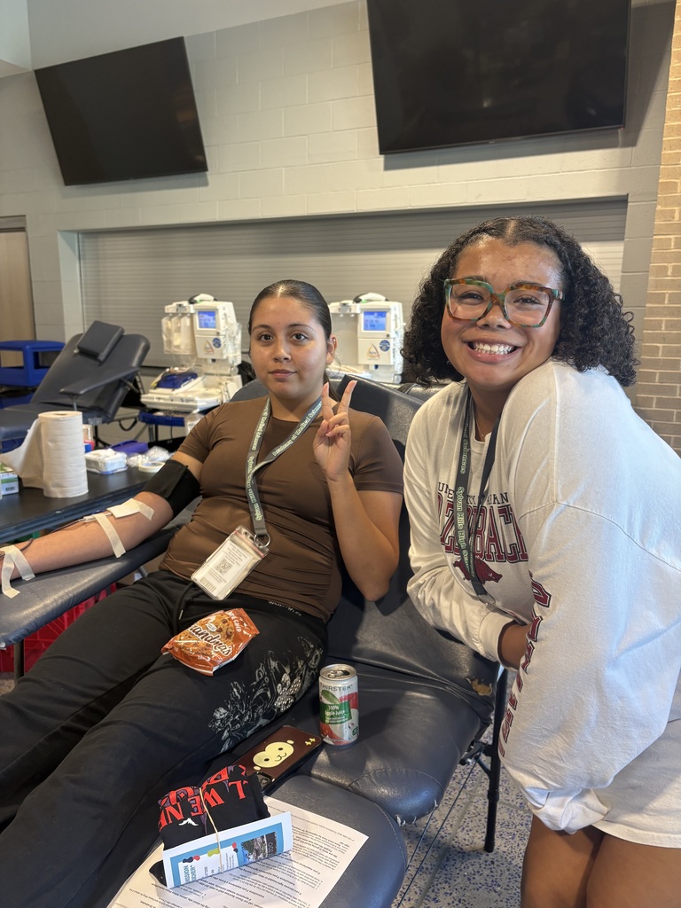 Two individuals stand in a blood donation area. One reclines in a chair with a bandaged arm, holding pretzels, a soda, papers, and a phone. The other stands beside them smiling. Medical machines and a brick wall are in the background.