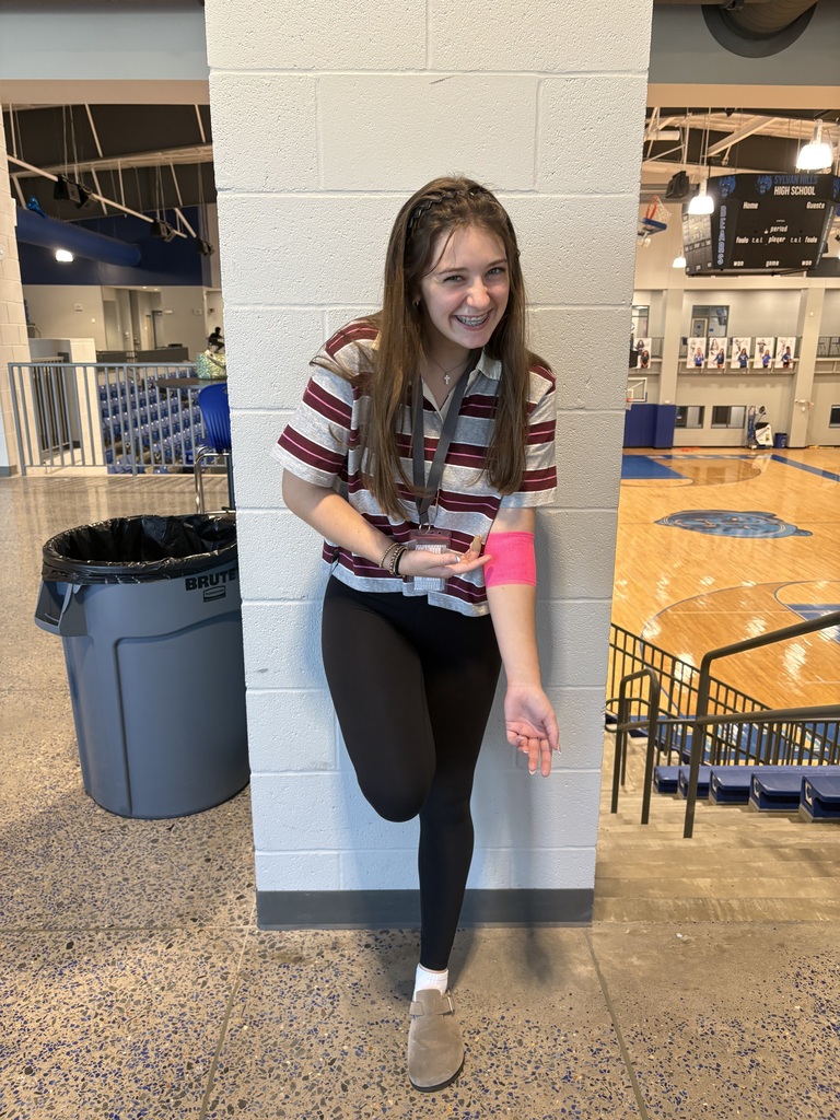 A person leans against a white brick wall inside a gym, wearing a maroon striped shirt and black leggings. A bright pink cast or bandage is on their right forearm. A trash bin, seating, and a basketball court are visible behind them.