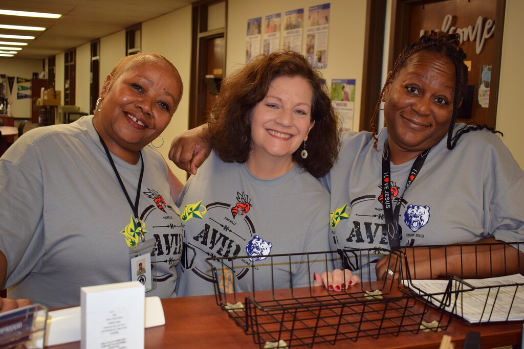 Three PCSSD Central Office staff members wearing grey AVID t-shirts smile together behind a reception counter.