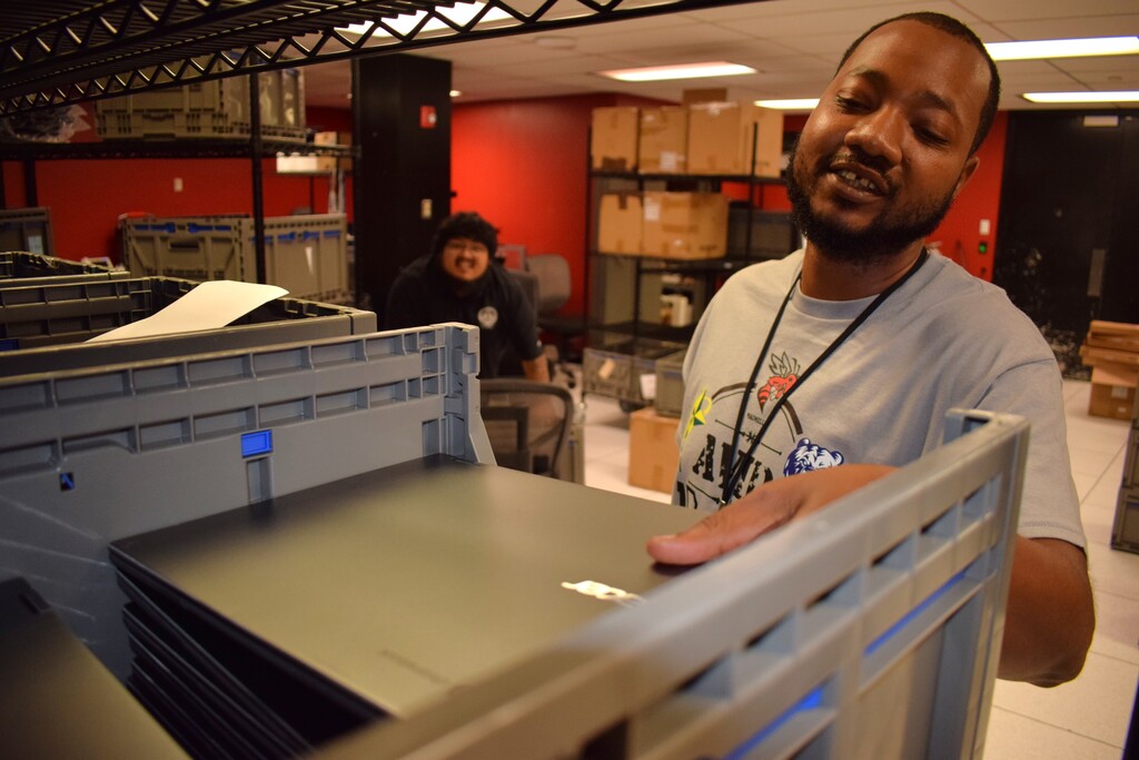 one personA PCSSD staff member wearing a grey AVID t-shirt reaches into a storage crate filled with technology equipment inside a workroom.