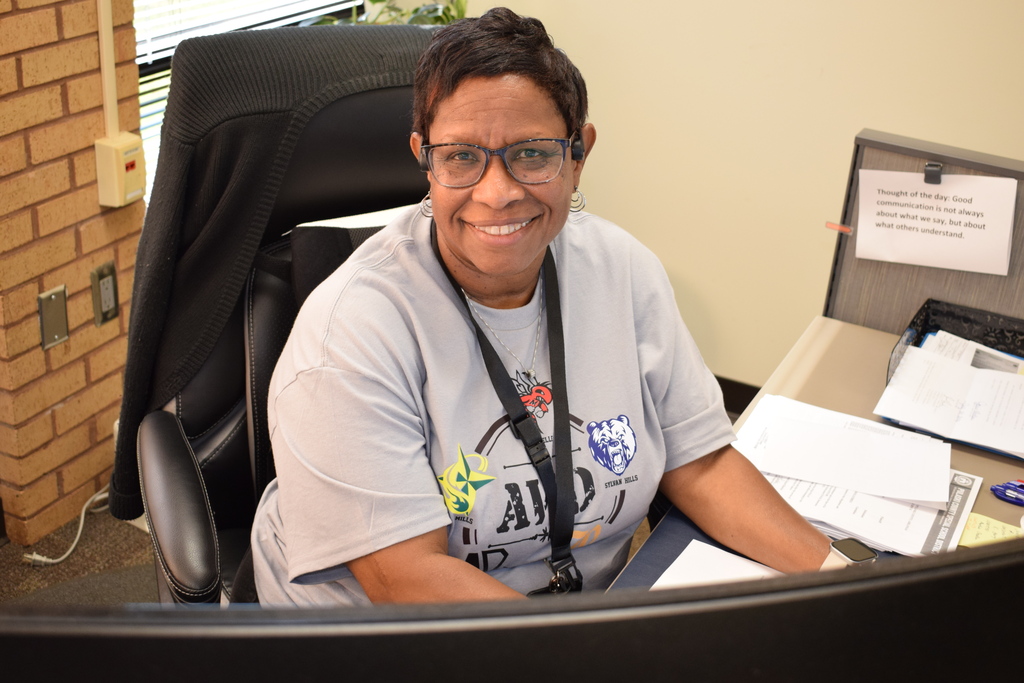 A PCSSD staff member wearing glasses and a grey AVID t-shirt smiles directly at the camera from behind her office desk.