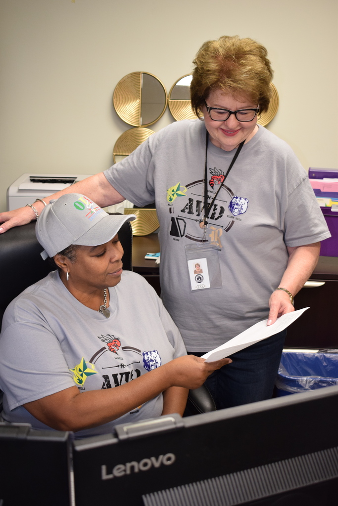 Two staff members wearing matching grey AVID t-shirts review a document together at an office desk.