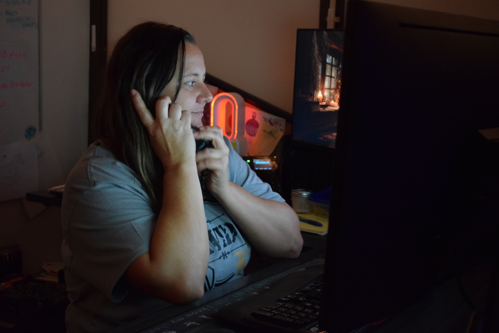 A staff member wearing a grey AVID t-shirt speaks on the phone while working at her desk in front of a computer screen.