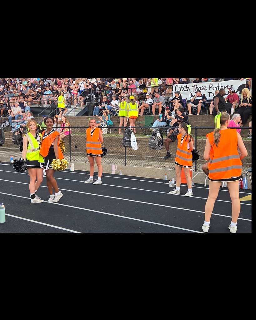 High school cheerleaders wearing bright neon orange and yellow safety vests over their uniforms pose on a black running track. Behind them, filled stadium bleachers display a banner that reads "Catch Those Panthers by the Tail."