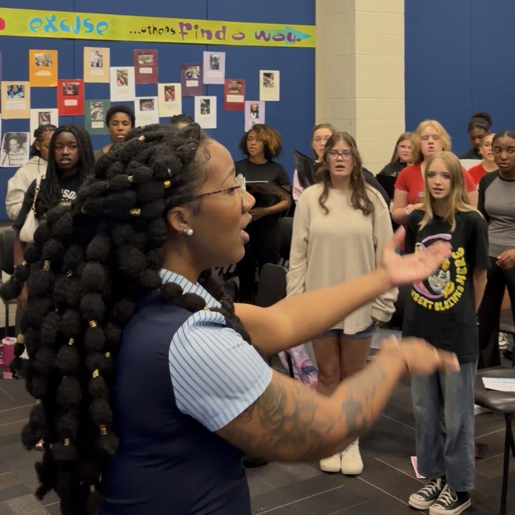 A choir stands in rows holding music folders while an instructor with beaded hair conducts at the front; colorful posters and photos cover the back wall.