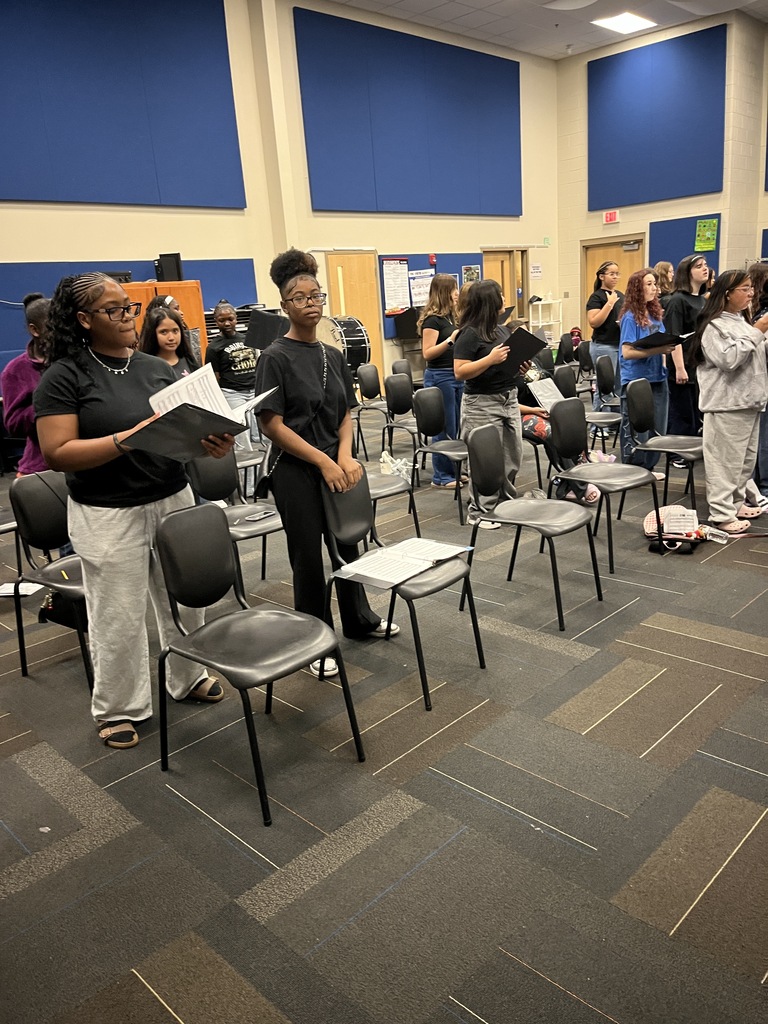A large group of high school choir students stand closely together in a music room, smiling for a group photo; blue acoustic panels and chairs surround them.
