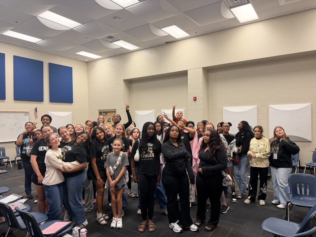 Students stand among rows of black chairs holding papers or folders in a music rehearsal room with blue acoustic panels and a drum set in the back.