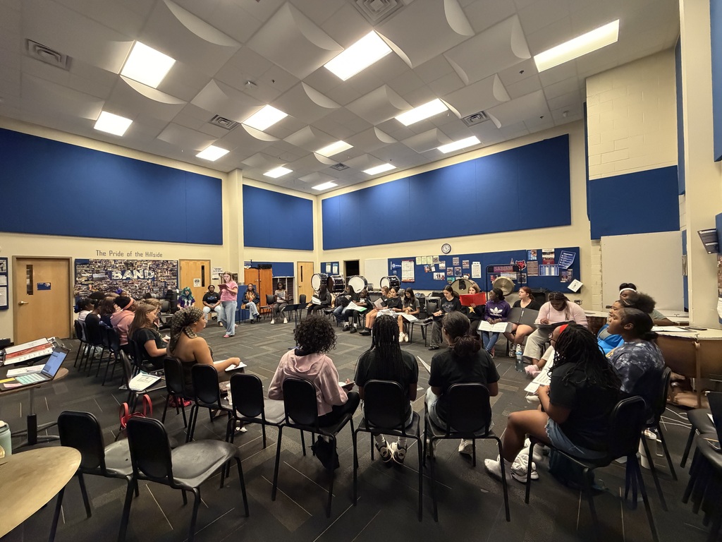 A group of students sit in a large circle of chairs in a band room while an instructor stands and leads discussion; percussion instruments and “The Pride of the Hillside Band” mural are visible in the background.