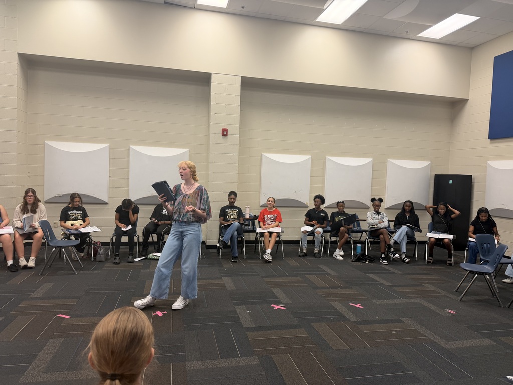 Students sit in a large circle of chairs while one student stands in the center reading from a tablet; the room has beige walls, acoustic panels, and pink tape X marks on the carpet.