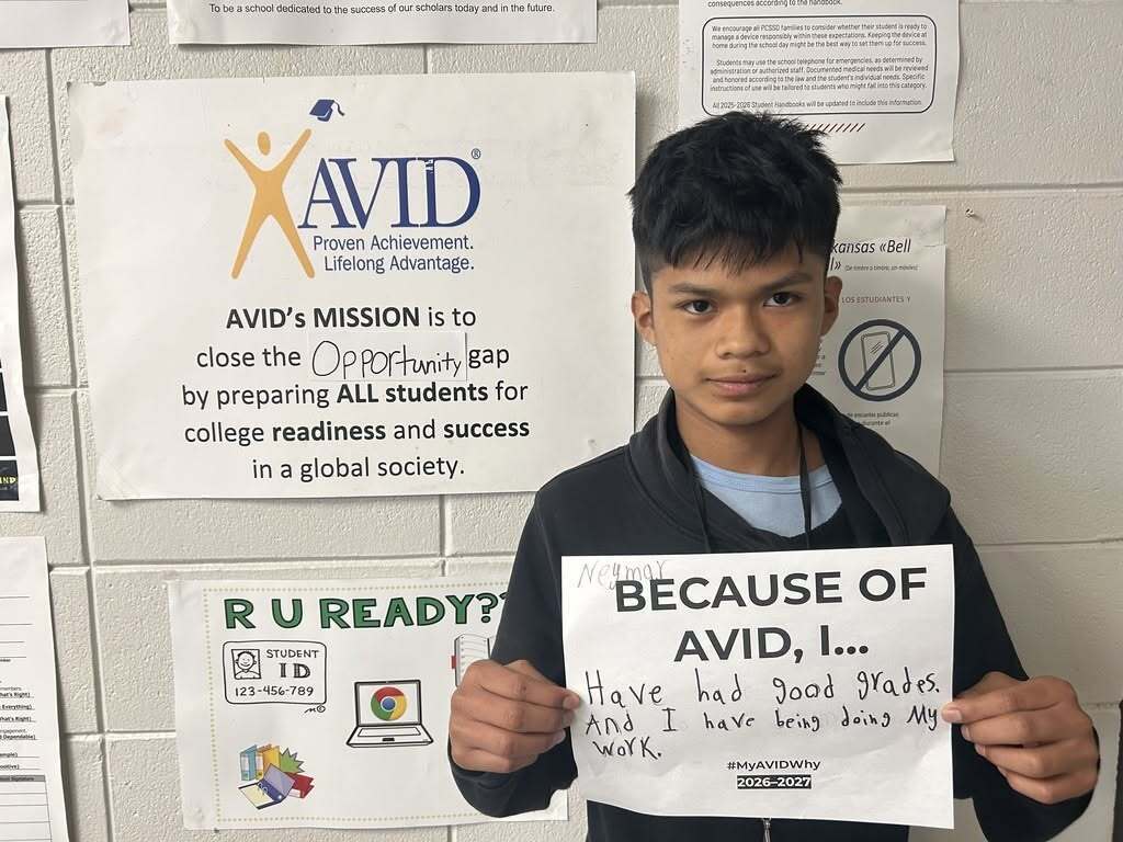 A student stands beside an AVID mission poster while holding a "Because of AVID, I..." reflection sheet. The handwritten response shares that AVID has helped the student earn good grades and stay focused on schoolwork. Surrounding wall displays include AVID materials and school information posters that reinforce college and career readiness.