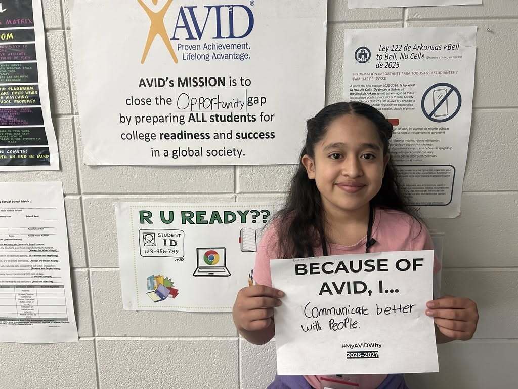 A student stands in front of an AVID mission poster and holds a "Because of AVID, I..." reflection sheet. The handwritten response reads, "Communicate better with people." Additional school posters about student readiness and school policies are displayed on the wall behind the student.