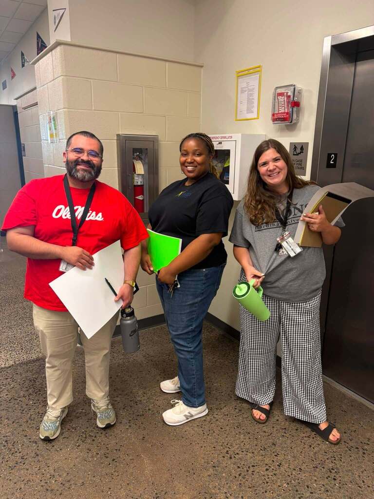 Three staff members stand together in a school hallway near an elevator, holding clipboards, folders, and other materials. The group is participating in an AVID activity and is surrounded by school safety equipment and hallway displays. The photo highlights staff engagement and support for schoolwide AVID initiatives.