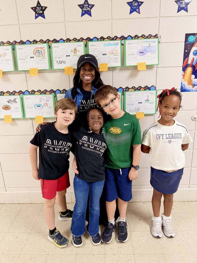Four elementary students and one staff member pose in a school hallway in front of a display of student work. The wall features vocabulary posters, space-themed decorations, and motivational star signs with encouraging messages. The group stands together beneath the student work showcase, highlighting a positive learning environment and student achievement.