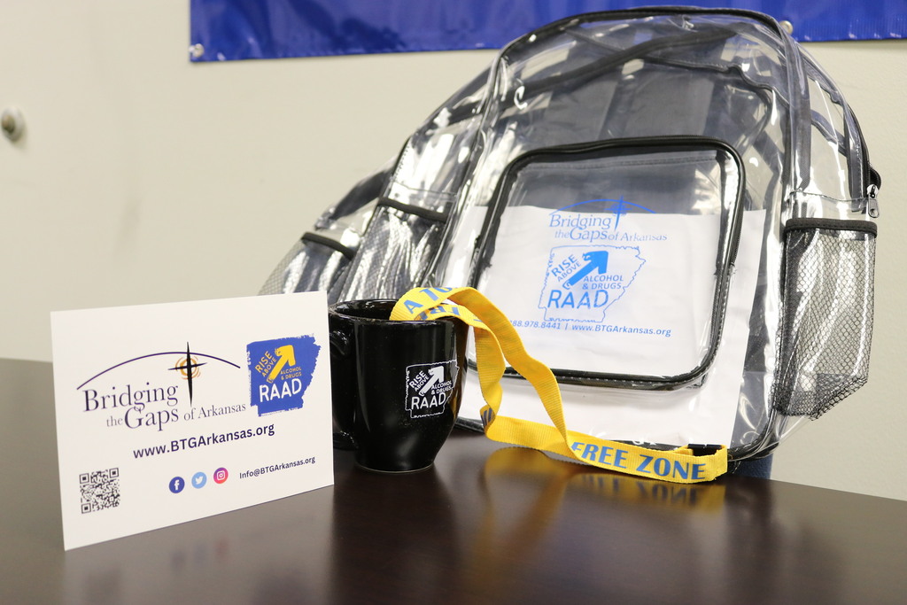 a clear backpack, a lanyard, a coffee cup, and a paper with Bridging the Gap of Arkansas branding on a table