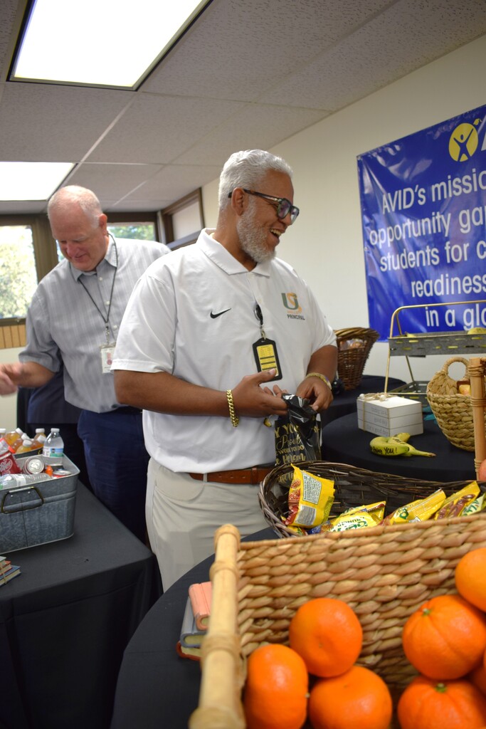 A principal with gray hair and glasses smiling as he picks out snacks from a table display. In the foreground, woven baskets are filled with fresh oranges and bagged snacks, with an AVID banner hanging in the background.