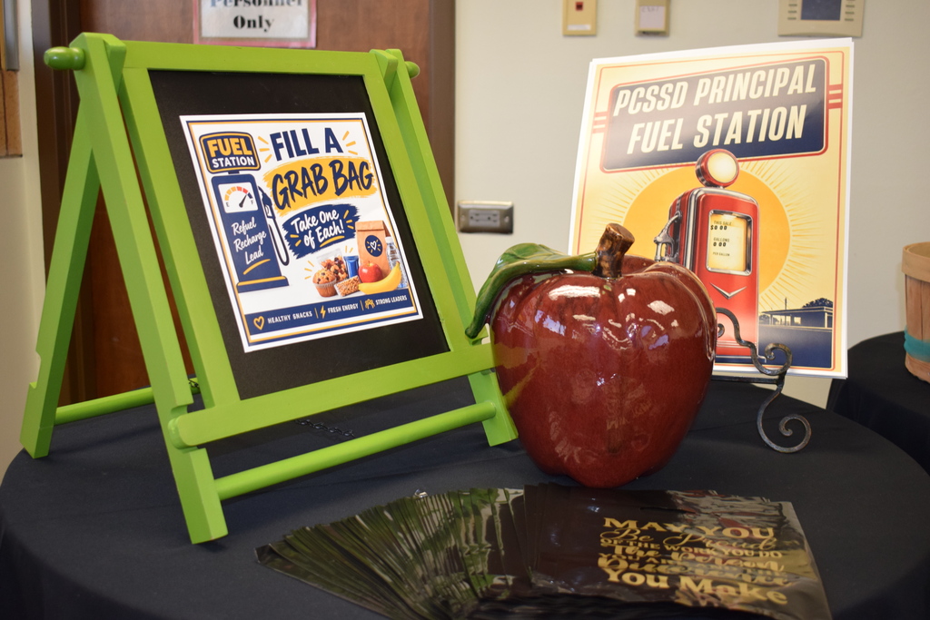 A tabletop display for the "PCSSD Principal Fuel Station." It includes a framed green sign reading "Fill a Grab Bag," a large ceramic red apple, black treat bags, and a poster featuring a retro gas pump labeled "PCSSD Principal Fuel Station."