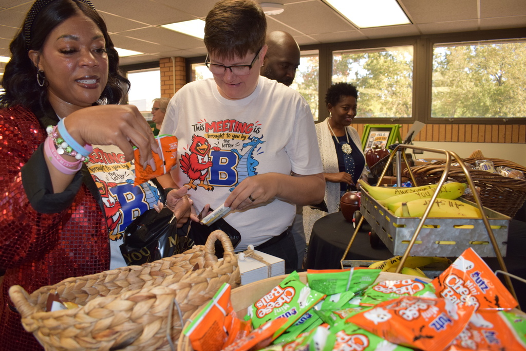 A group of school leaders browsing snacks at the fuel station table. In the foreground, a woman in a red sequined blazer grabs a snack bag, while another person wears a t-shirt that says "This MEETING is brought to you by the letter B." Baskets of snacks and fresh bananas sit on the table nearby.