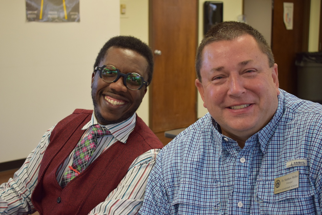 Two male principals sitting next to each other at a table, smiling at the camera. The principal on the left wears a maroon vest, plaid tie, striped shirt, and dark-rimmed glasses. The principal on the right wears a blue plaid button-down shirt with a name badge.