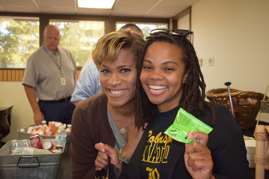Two female school leaders smiling together at the fuel station. One holds a small green snack pack, while beverage tubs filled with ice, canned drinks, and water bottles sit in the background.