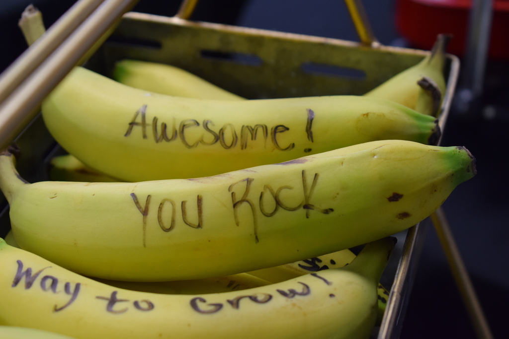 A close-up view of fresh yellow bananas stacked in a wire basket, with encouraging handwritten messages written on their peels in black marker, including "Awesome!", "You Rock", and "Way to Grow!"