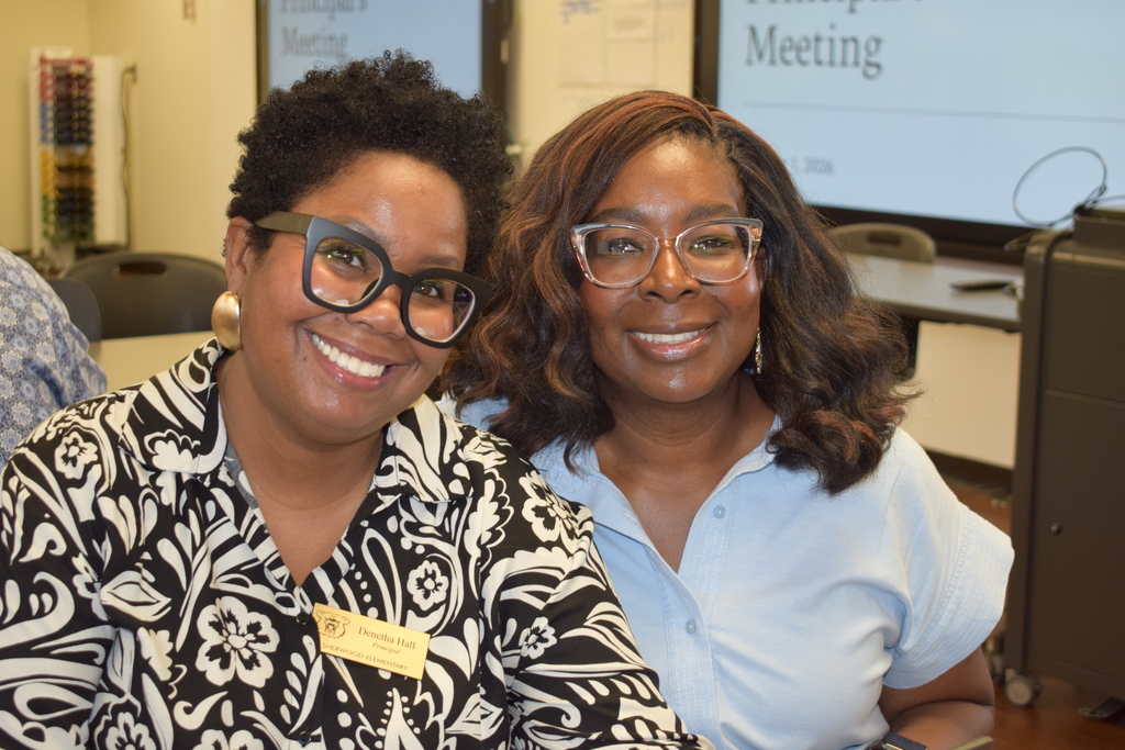 Two female principals smiling together indoors in front of a presentation screen reading "Principal's Meeting". The woman on the left wears black-framed glasses, large gold earrings, and a black-and-white patterned top with a name badge. The woman on the right wears glasses and a light blue top.