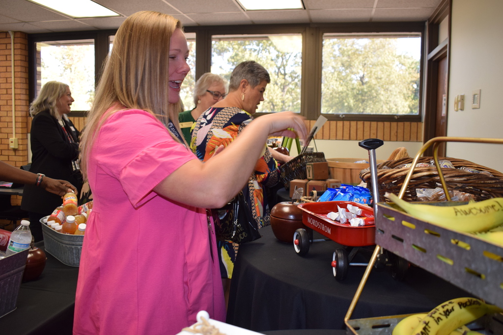 A woman in a bright pink dress smiling as she reaches into a snack basket at a decorated table. In the foreground sits a small red toy wagon filled with wrapped treats alongside a basket of bananas.